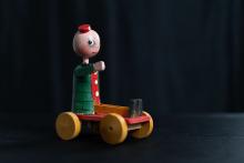 Comicfigur Fortunello als gedrechseltes und bemaltes Spielzeug sitzend auf einem Räderbrett, der beim Schieben die Beine auf und ab bewegt